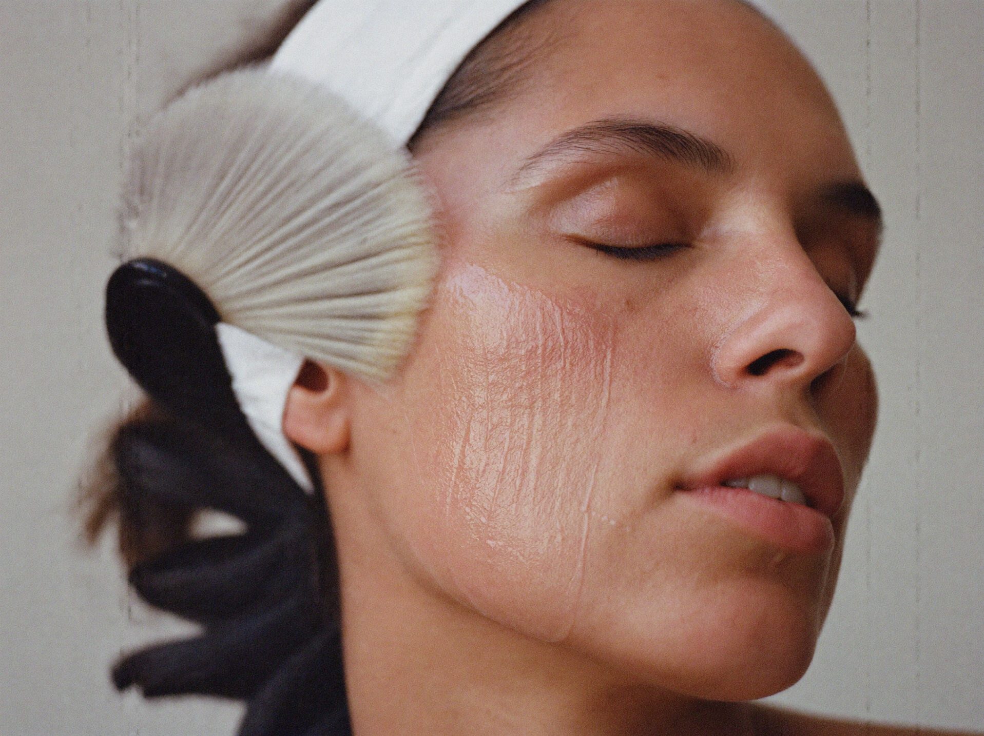 A fan brush sweeping a peel across a woman's cheek