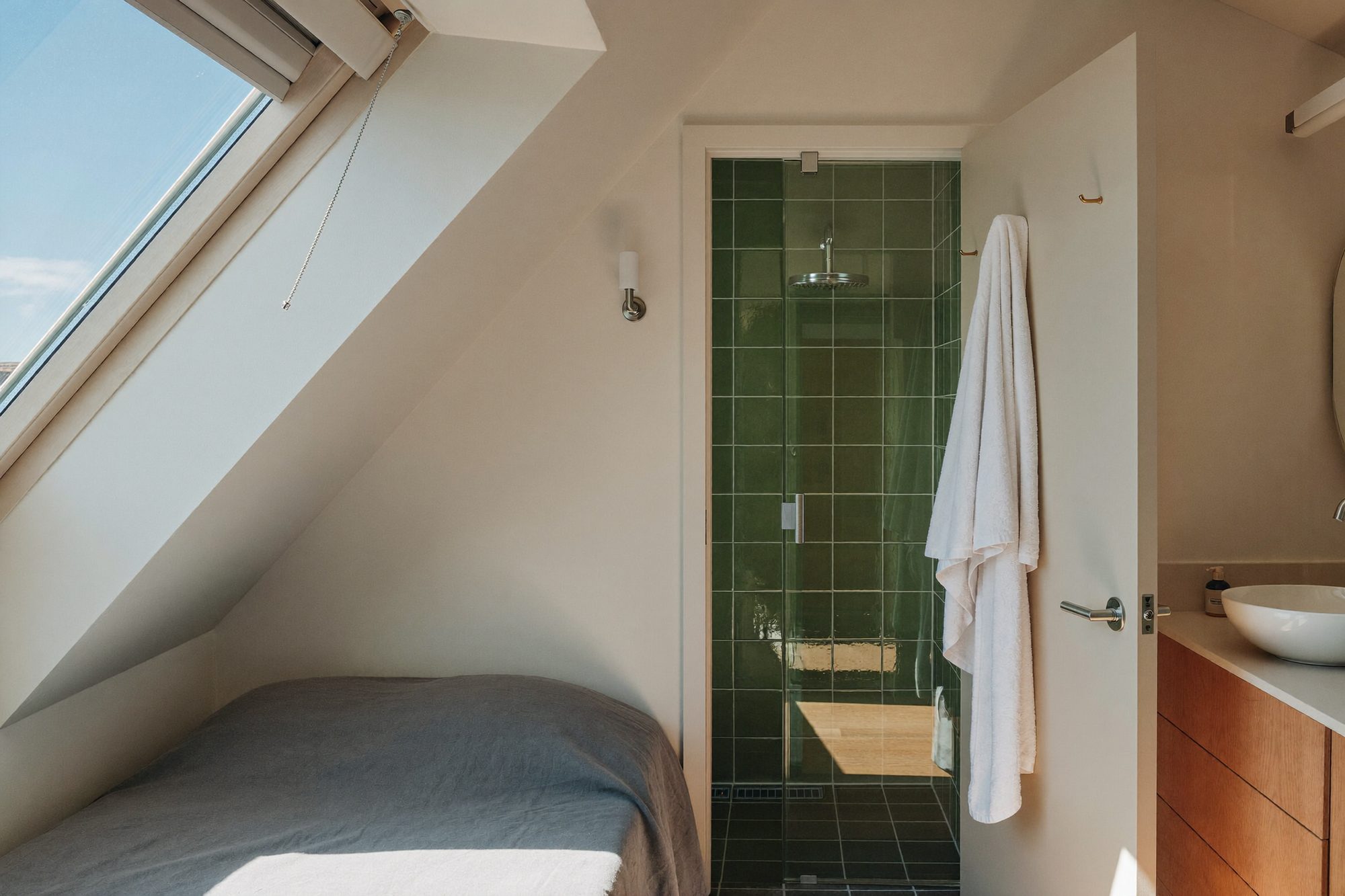A loft bedroom under a sloping ceiling with a green-tiled shower room through the door